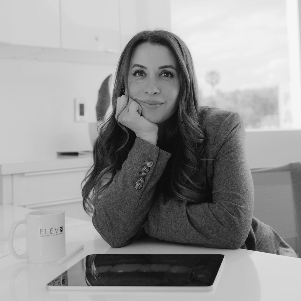 Dr. Leedia Riman at her desk at ELEV8 in Beverly Hills, chin resting on her hand, with a tablet and an ELEV8 mug in front of her